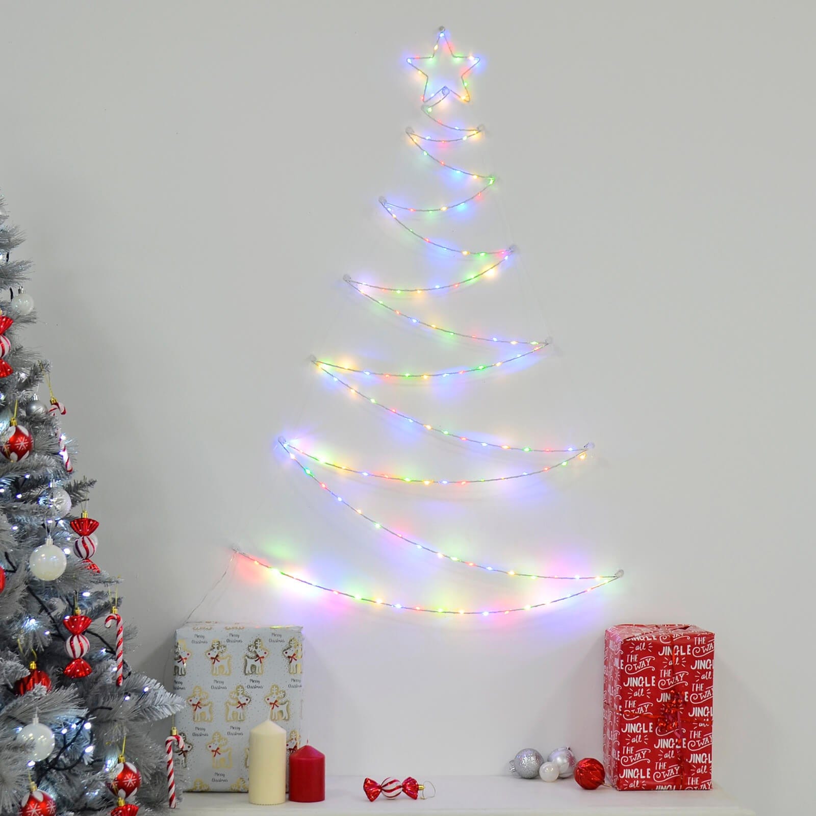 Light up Christmas tree shape festive wall hanging decoration with multicoloured LED lights beside a silver Christmas tree with red baubles, above a table with presents, candles and decorations